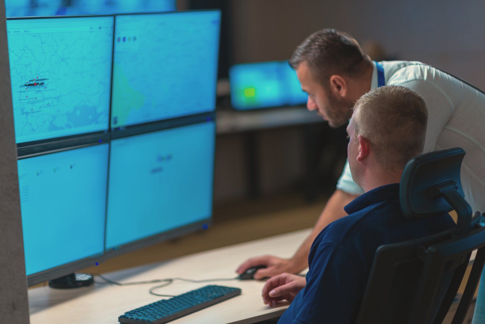 An engineer leaning in to walk a seated analyst through what's on their security monitors