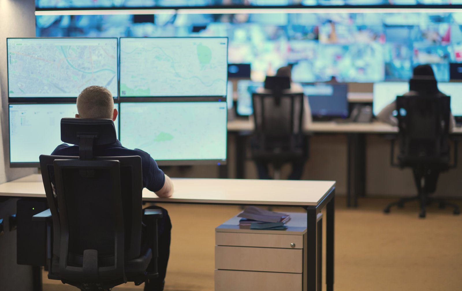 An analyst at a multi-monitor workstation in a security operations center, with a wall of monitoring screens behind
