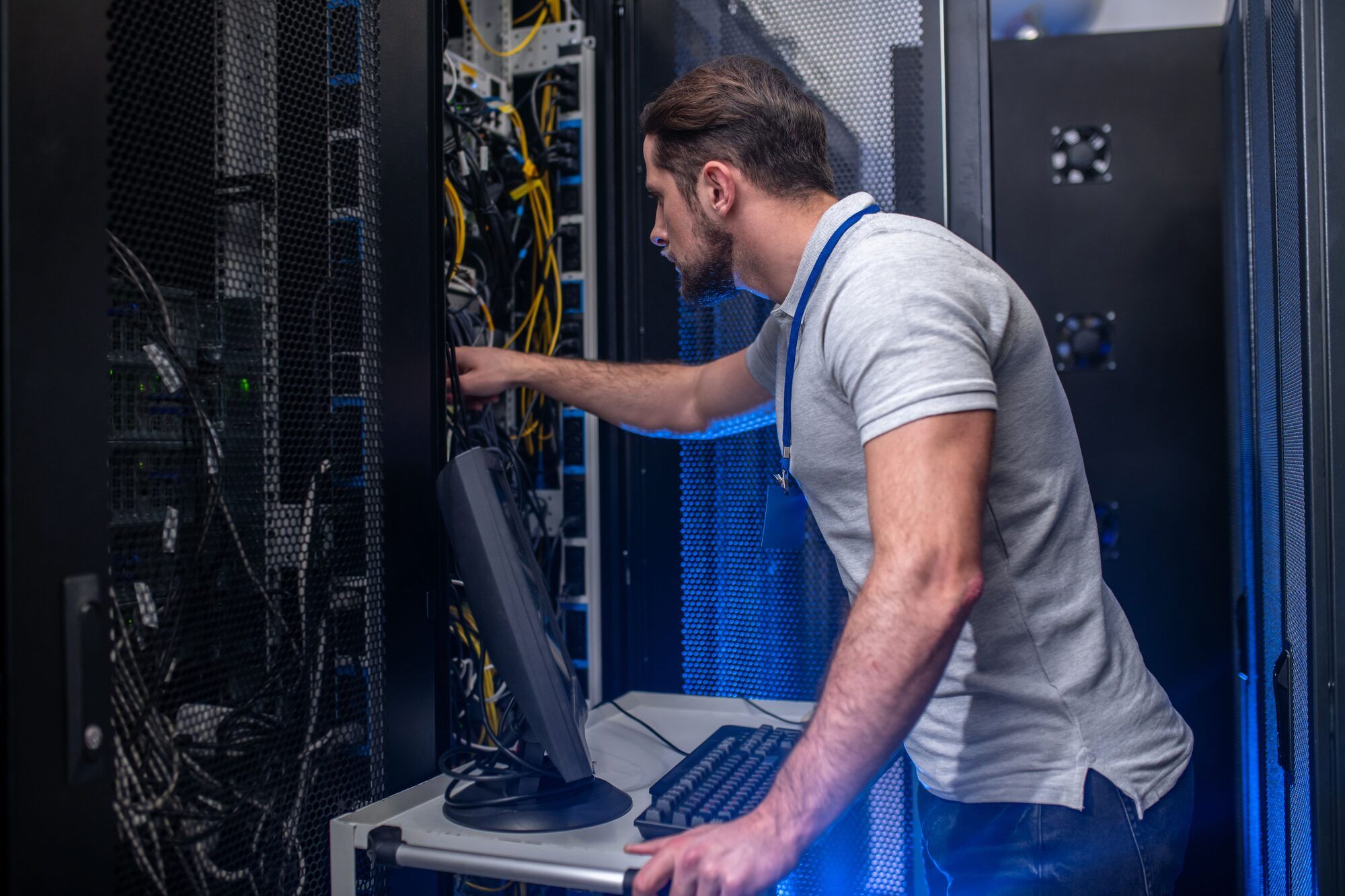 A technician working at an open server rack, one hand on the cabling and a monitor and keyboard on a cart beside him